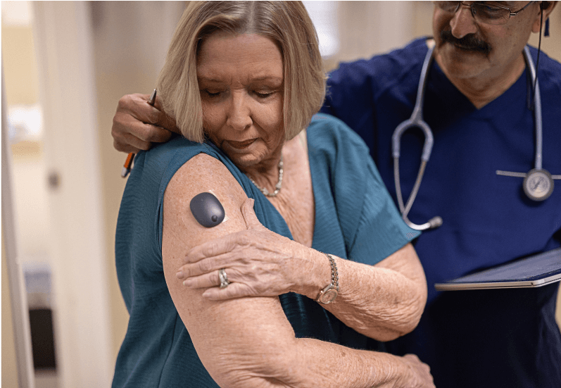 Woman adjusts device on arm while doctor observes in medical office, wearing teal shirt and he holds a clipboard.