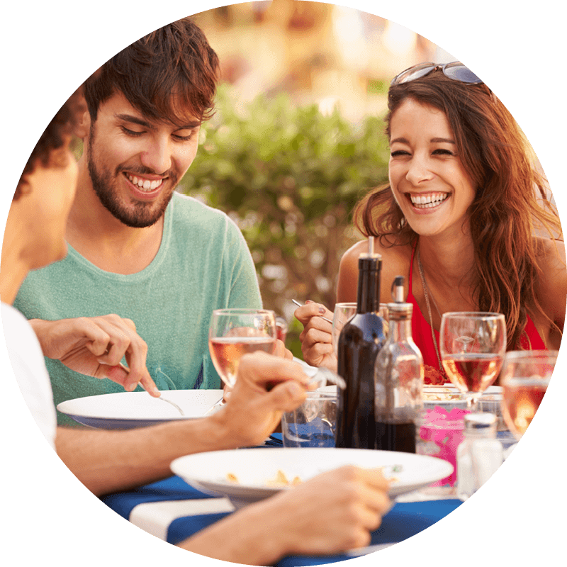 Three people smile and eat together at a table outdoors, surrounded by drinks, bottles, and blurred greenery.